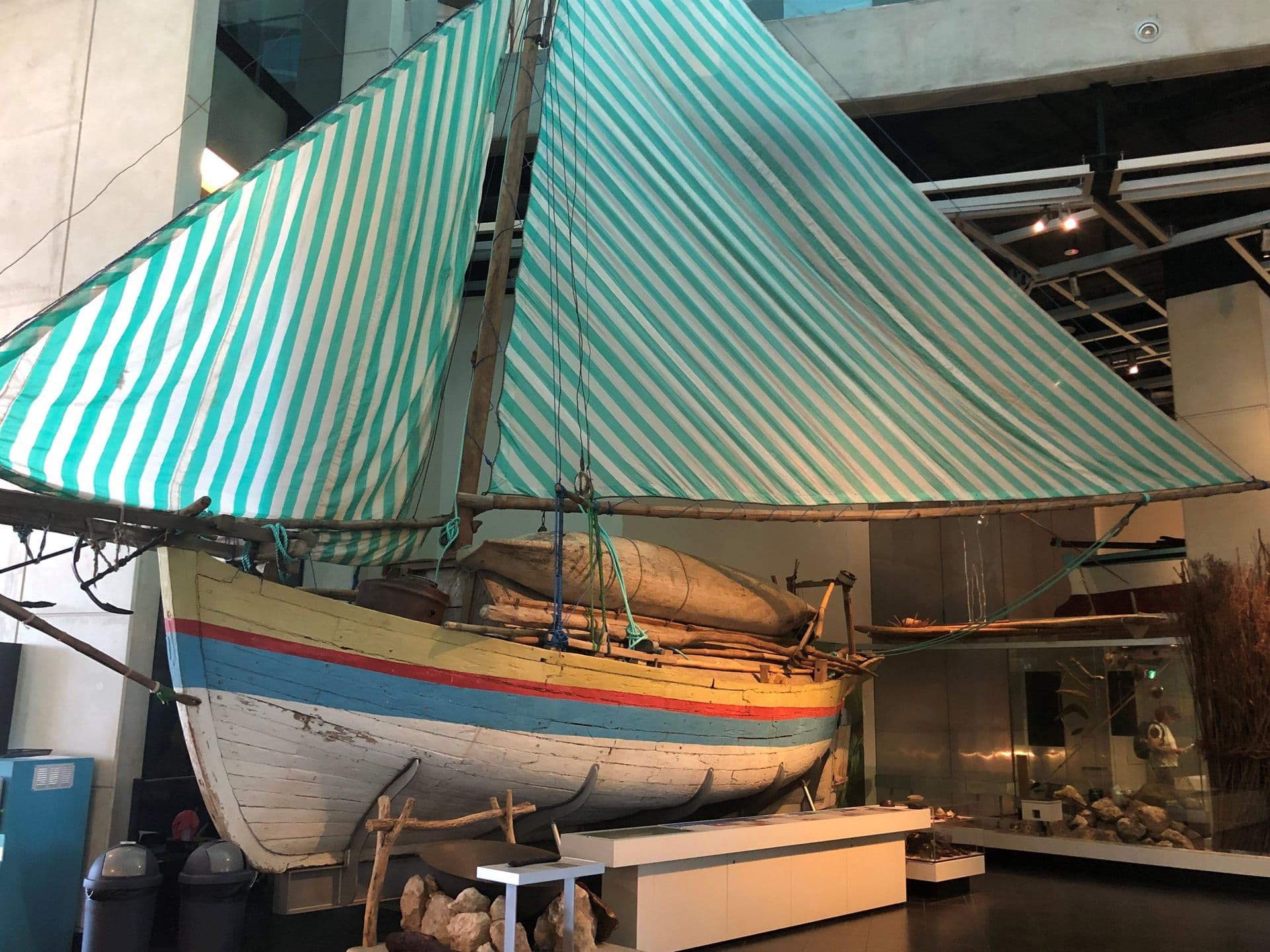 A small wooden sailing boat. The hull is painted white with yellow, red and blue stripes. The sails are striped blue and white.