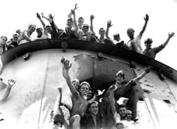 Many men, some wearing white sailor's caps, wave from the top of a ship's funnel. A puncture hole in the side large enough to fit four people alongside is also crowded with men waving.