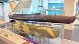 Two large canoes mounted behind a glass shield. One is higher up and covered in black canvas, the lower one is metal painted light and dark brown.