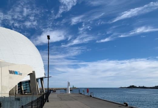 A view of the ocean and blue sky with streaks of cloud taken from the dock of the harbour. The white building of the WA Maritime Museum with its curved roof is on the left of the image and the mouth of the river heading out to the ocean is at the centre.