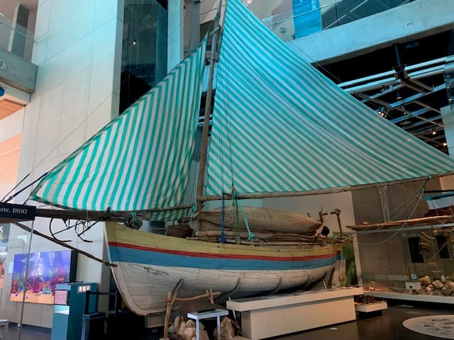 A small wooden sailing boat. The hull is painted white with yellow, red and blue stripes. The sails are striped blue and white.