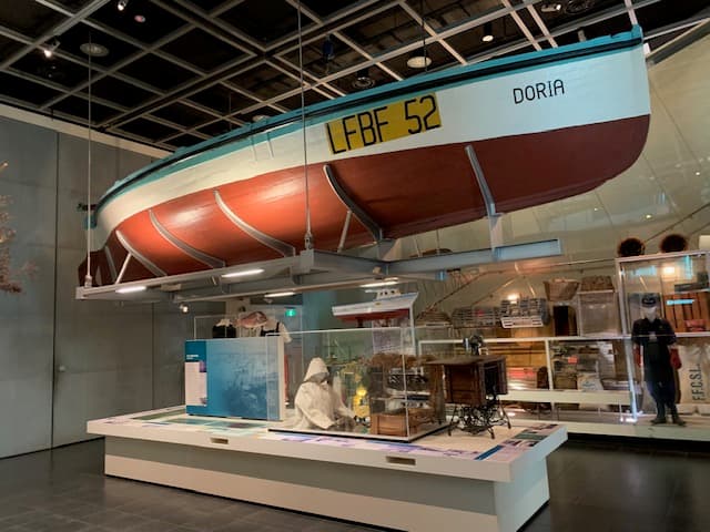A small wooden boat with the name of Doria. Its bottom half is painted a dull red, the top half is white, and the rim of the boat is blue.