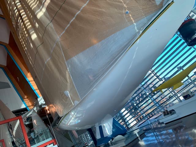 A large white yacht on display in a gallery. It has a fully extended tan-coloured sail, and its keel underneath the hull is white and blue.