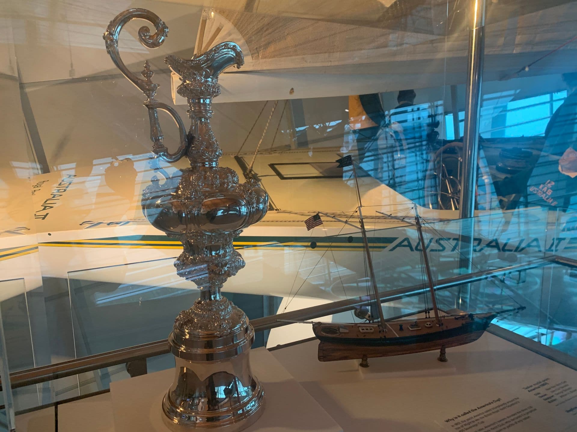 An ornamental silver jug, also called a ewer, in a display case. Behind can be seen the deck of a racing yacht with mannequins on board.