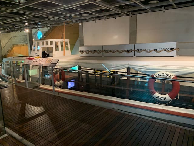 The Valdura ferry sits on display alongside a boardwalk with a glass balustrade. A life preserver bearing its name is visible on the side of the boat.
