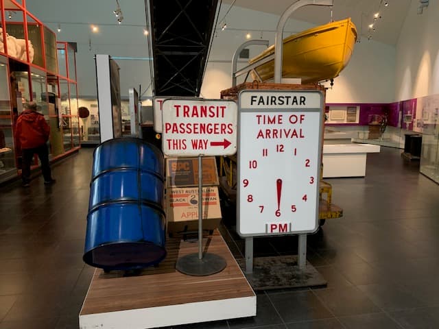 Alt text: A collection of metal signs are gathered in front of an old-style luggage trolley. One sign states ‘Transit Passengers this way’ and the other sign is a clock announcing the time of arrival of the Fairstar ship. There is a blue metal barrel and boxes displayed alongside.