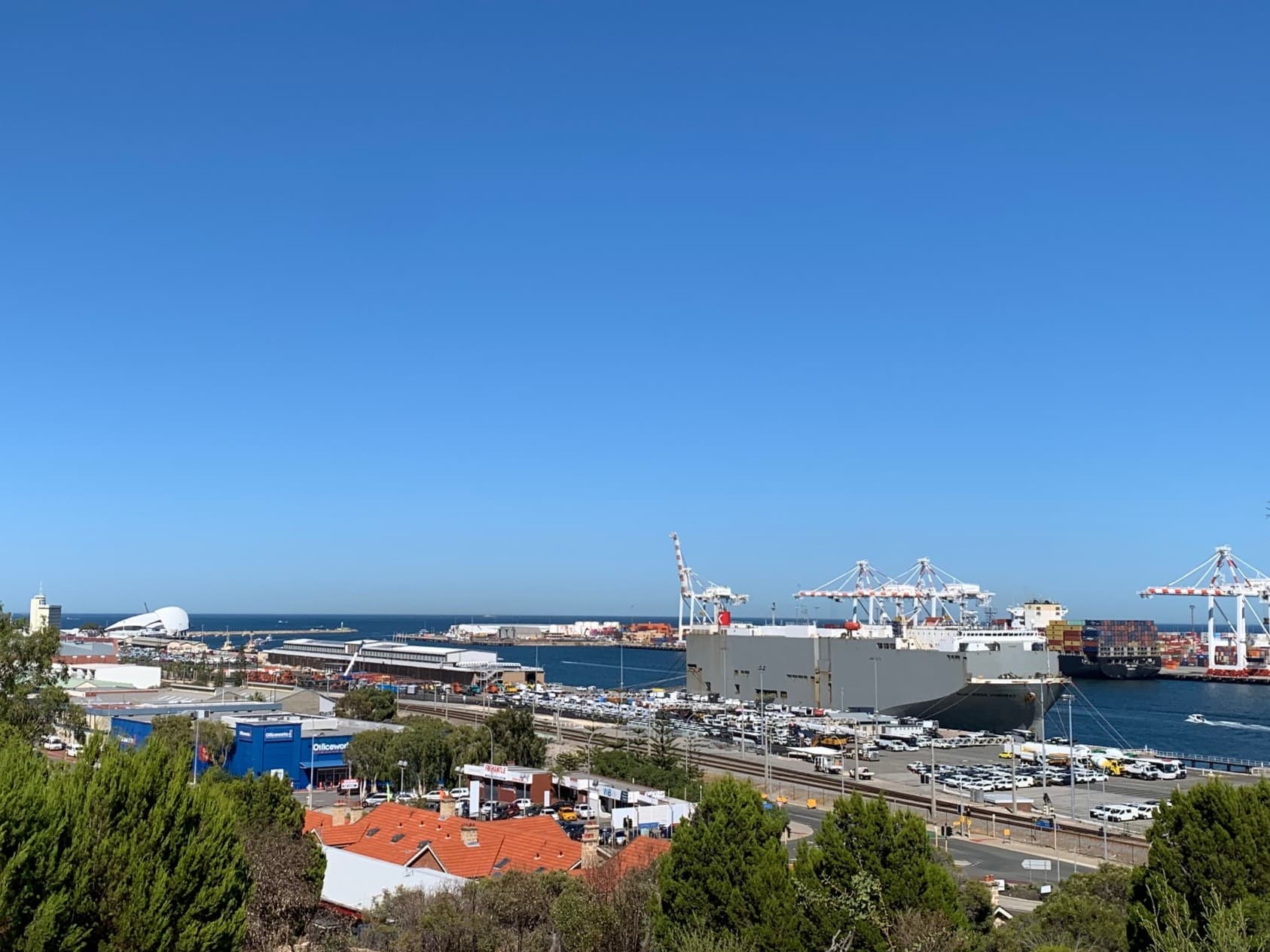 A view of Fremantle Port taken from Cantonment Hill. The sky is a clear bright blue and the water is a deep blue. Visible are some container ships, the iconic Port cranes and the WA Maritime Museum at the entrance to the port.