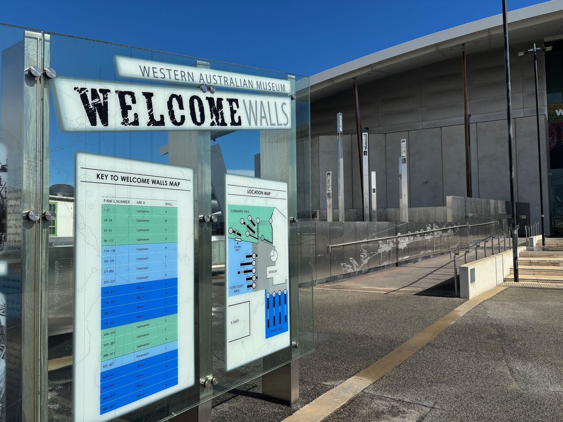 A glass sign with Welcome Walls and images is positioned in front of a building with a white curved roof and concrete walls.