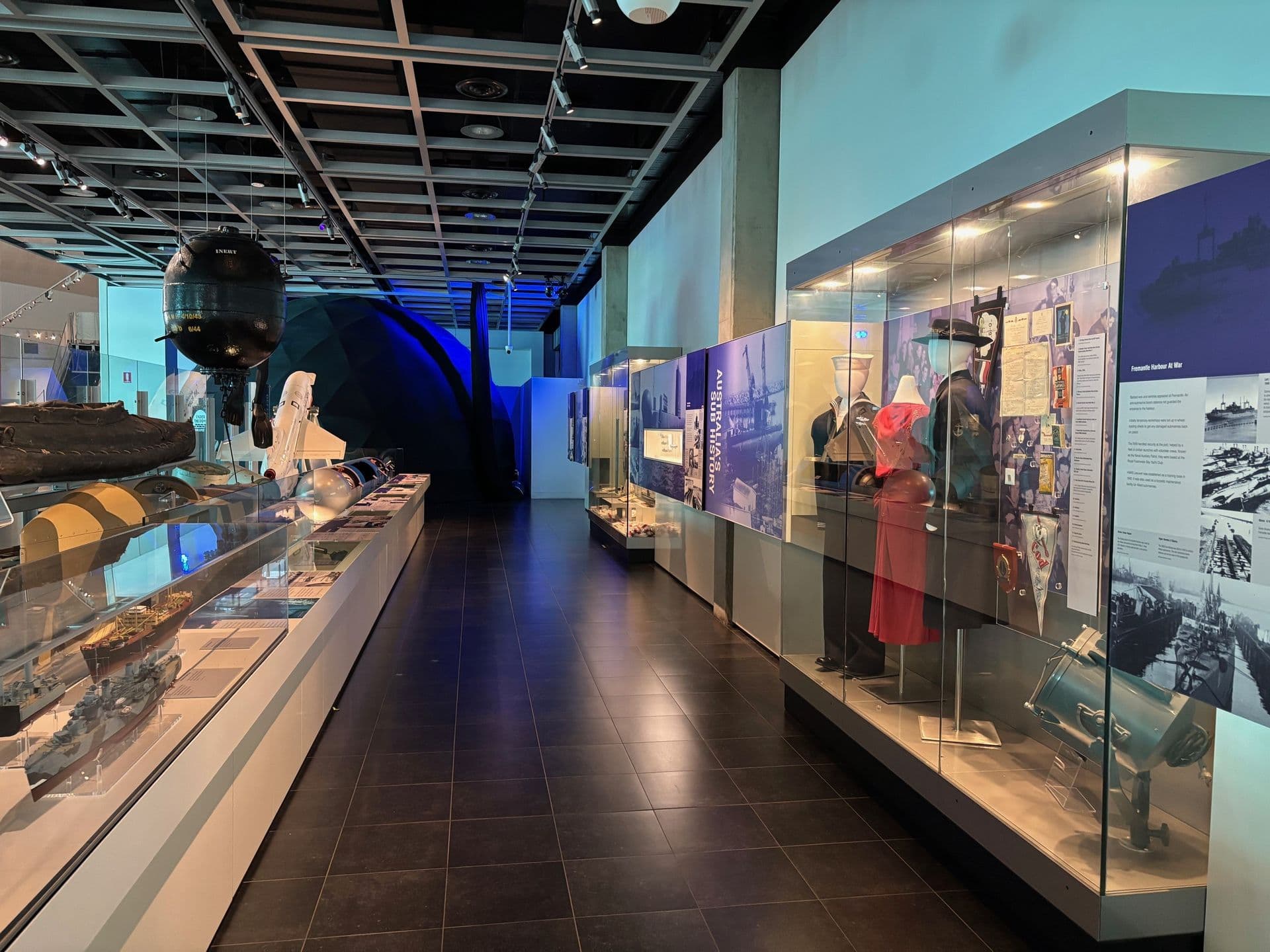 A walkway between gallery cabinets displaying war related items, including clothing and model ships.