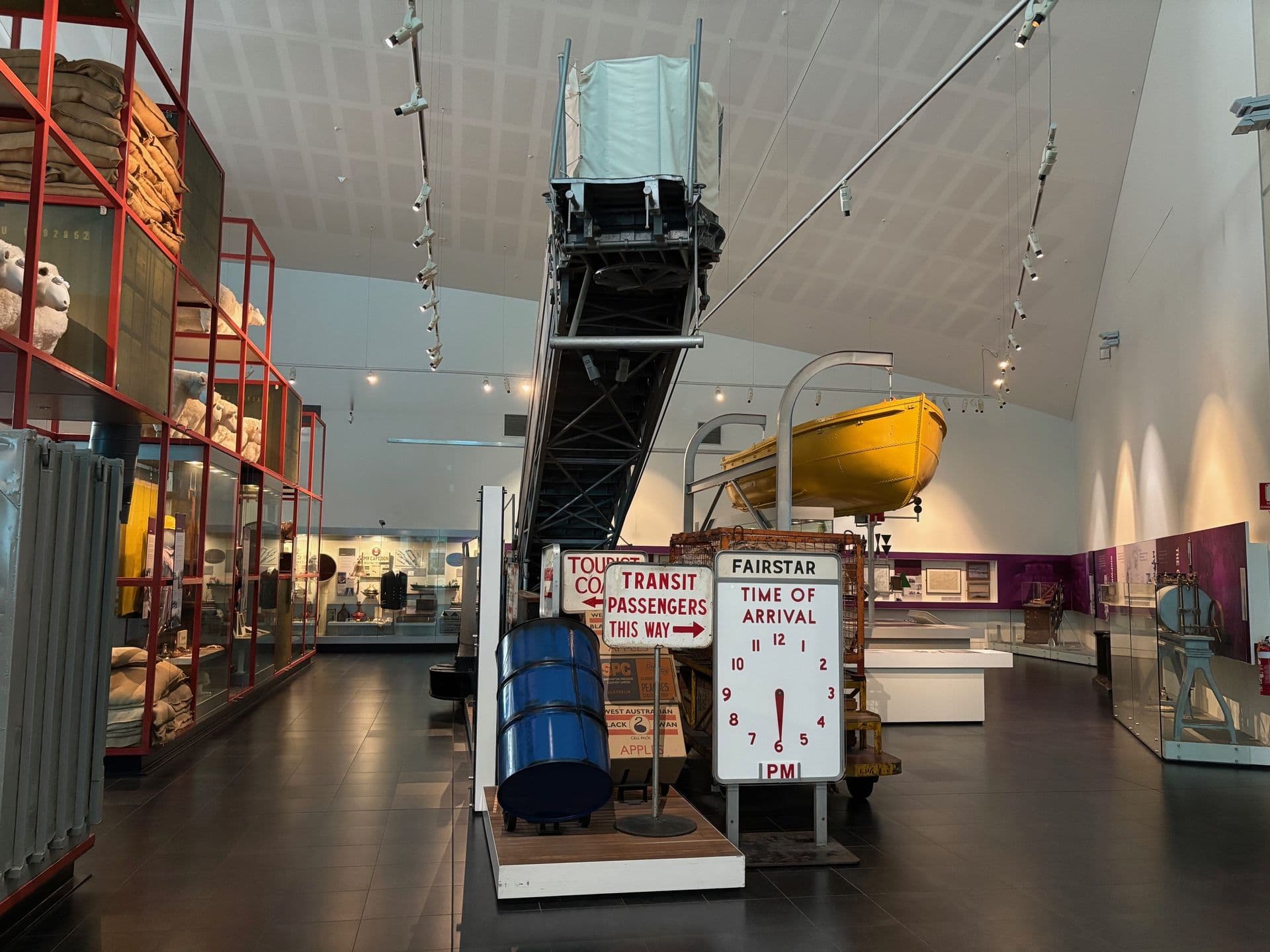 A ship's gangway stretches up high into the gallery. Underneath are some vintage passenger arrival signs displayed with cargo items on trollies, such as boxes and a blue metal barrel.