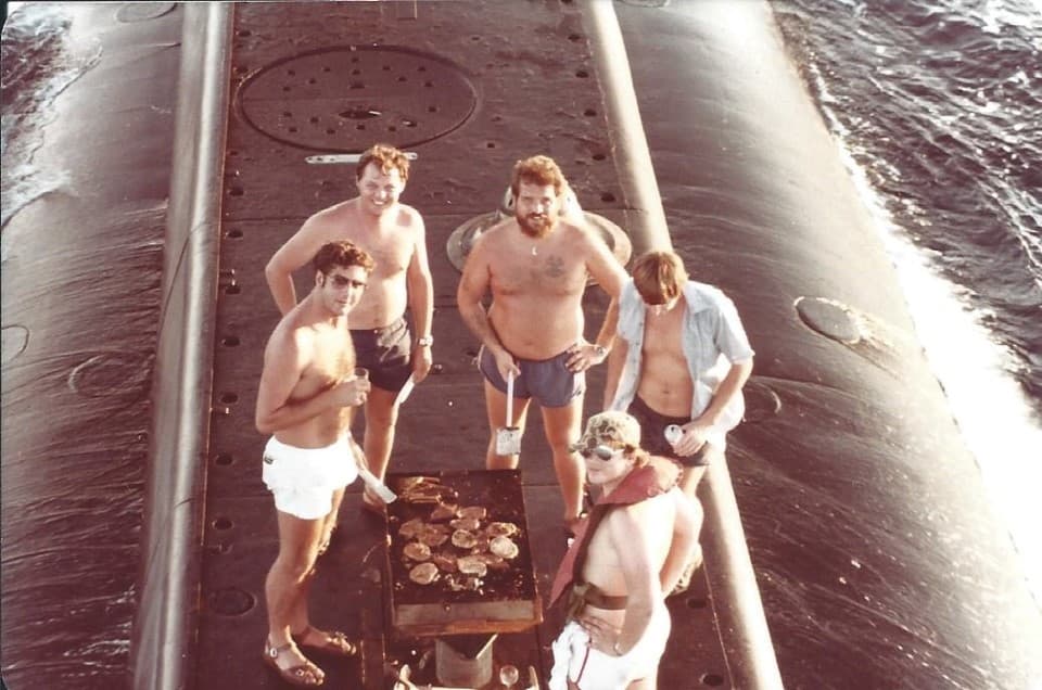 On the top of a submarine in the water stand five submarine crew, four of them dressed in only shorts, surround meat cooking on a barbecue.