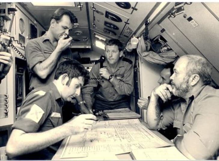 Submarine crew gathered around a table looking at charts. One is holding a telephone and another is speaking into a radio.