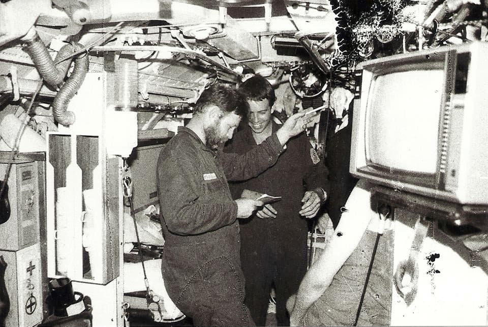 A grainy black and white photo of two crew in overalls working in the tight corridor of the submarine.