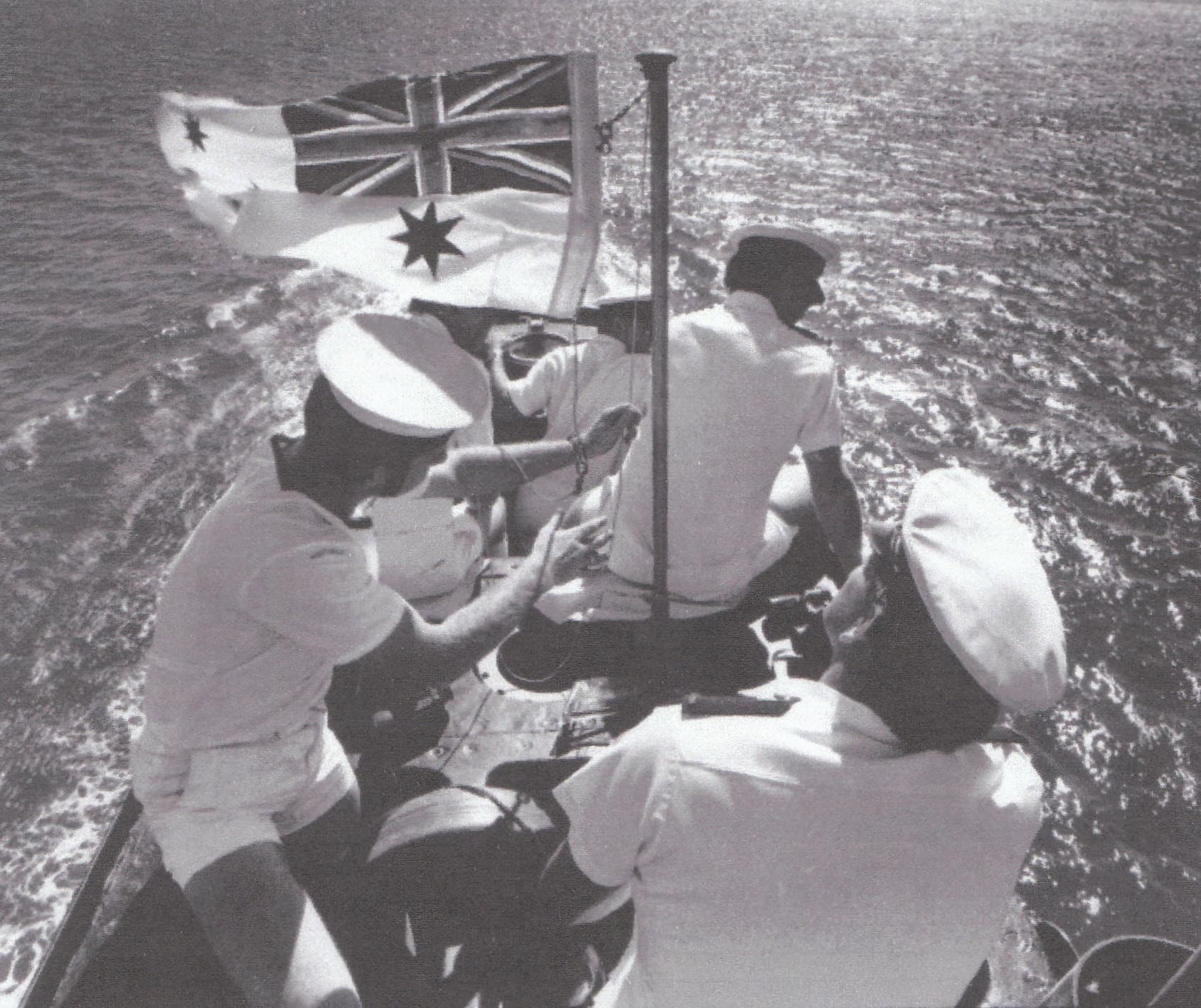 A black and white photo of several sailors in clean white uniforms gathered on top of the sub. An Australian flag is raised.