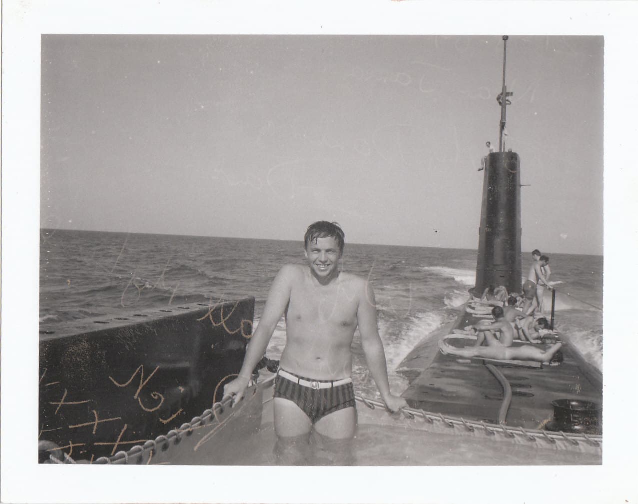 On the top of the submarine, a sailor in a bathing suit smiles from a fabric shallow pool filled with water. Several other men sun themselves on the deck.