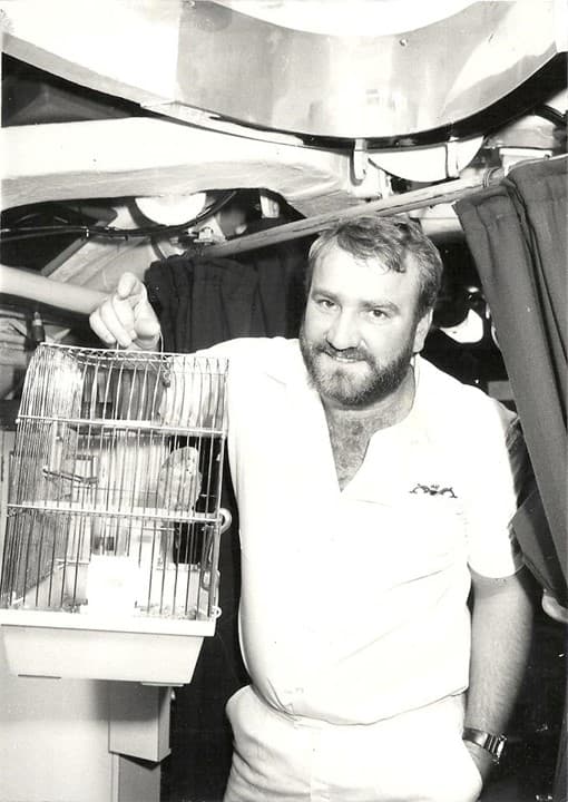 A grinning crewmember holding a small bird cage containing a budgerigar sitting on a perch.