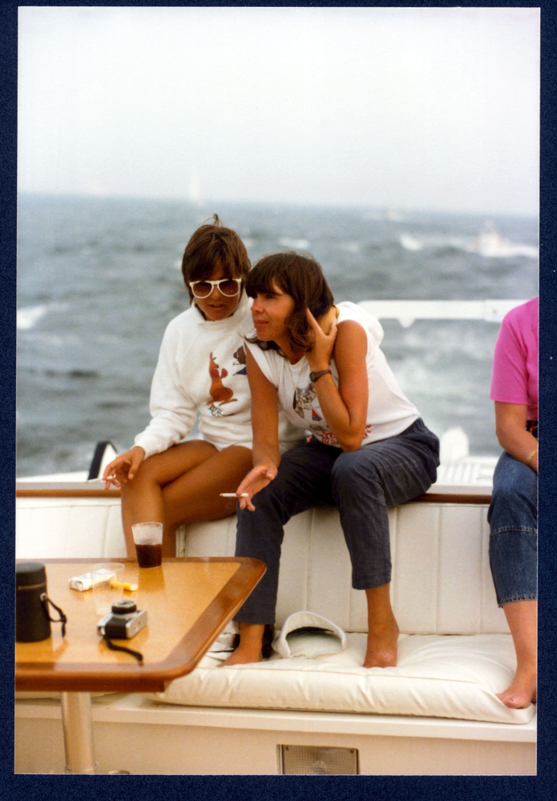 Two young women in 1980s clothing, both with cigarettes, are perched at the edge of a boat's deck. One has an arm around the other as they chat.