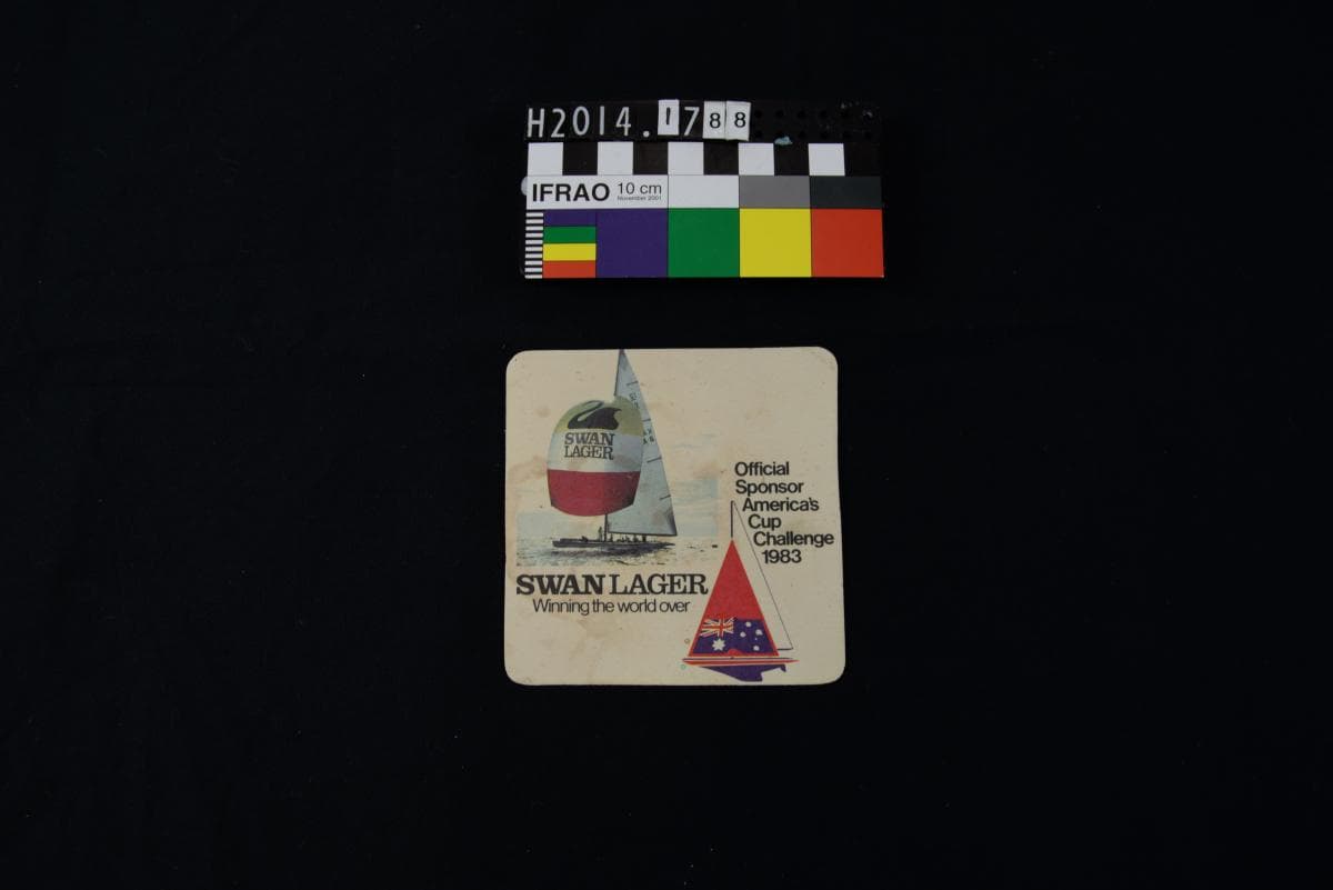 A white beer coaster, discoloured with age. A yacht is depicted. Text reads "Swan Lager: Winning the world over. Official sponsor, America's Cup Challenge 1983."