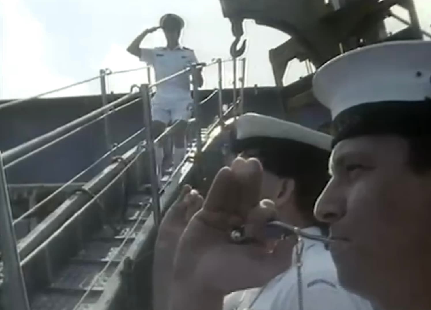 Two sailors blowing small musical instruments stand beside a walkway as a man in a white uniform and hat walks down it, saluting.