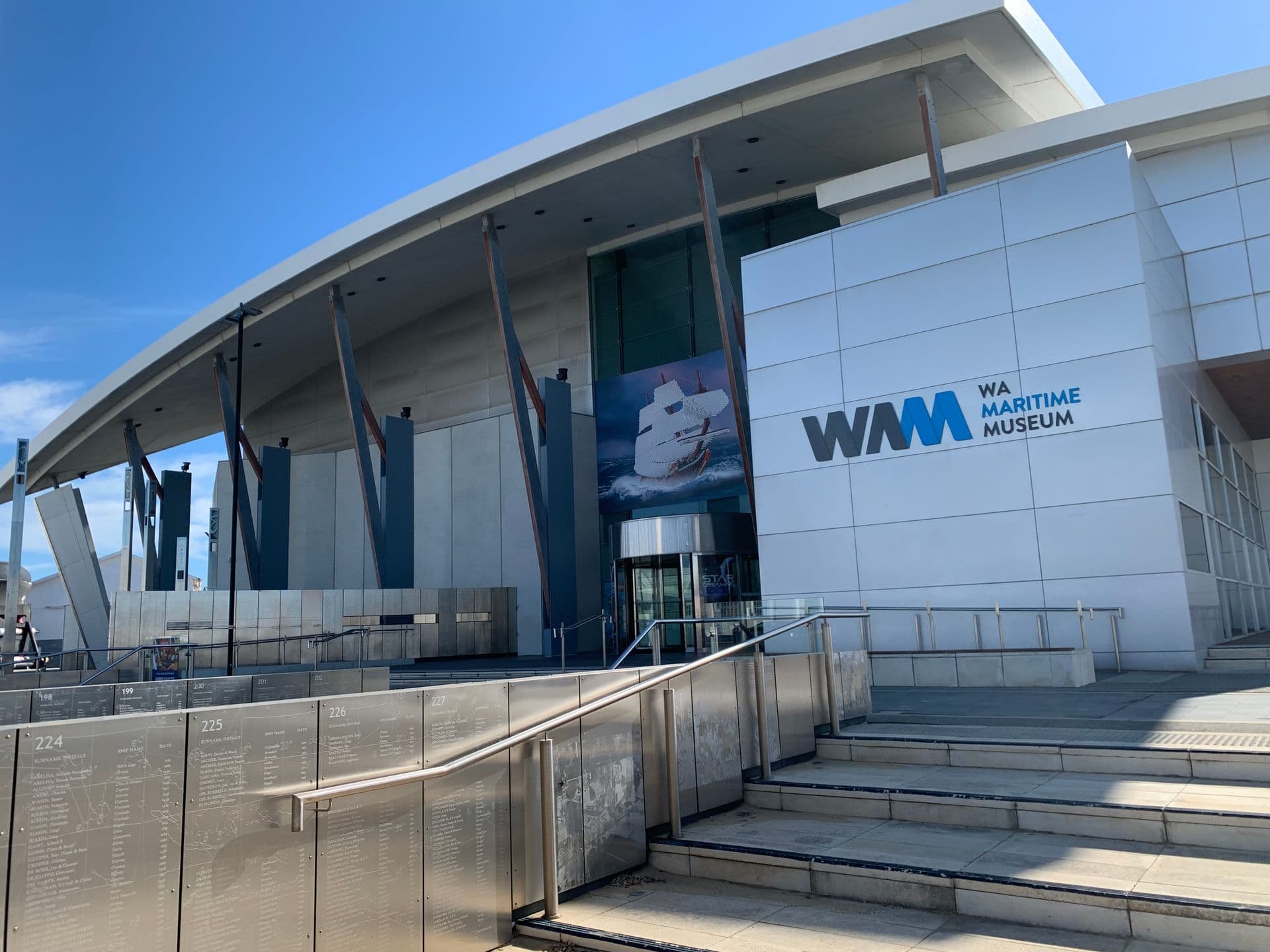 In the foreground are silver panels with writing on them. Steps lead up to a large white and grey building with the name WA Maritime Museum.