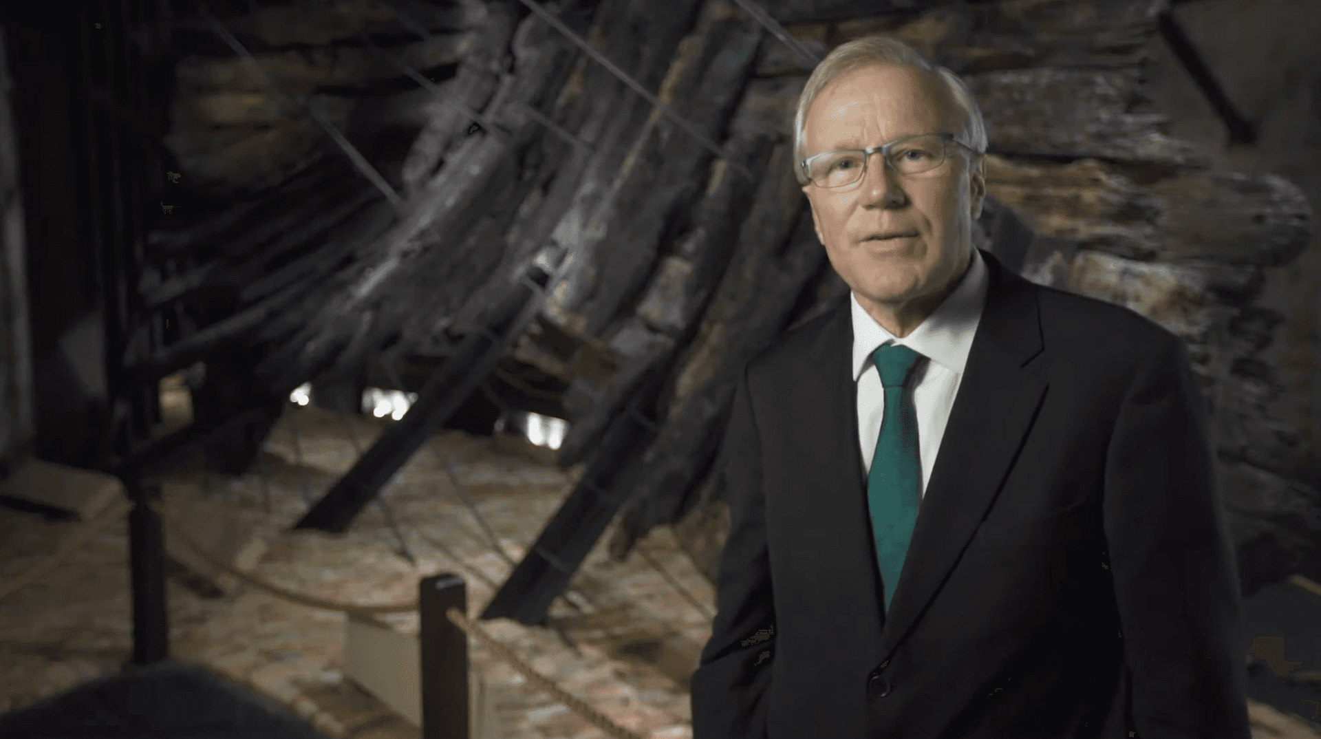 A man in a suit standing in front of the timbers of the Batavia shipwreck.