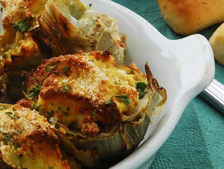 A cooked artichoke with breadcrumb topping browned in the oven sits in a white baking dish on a green table cloth.