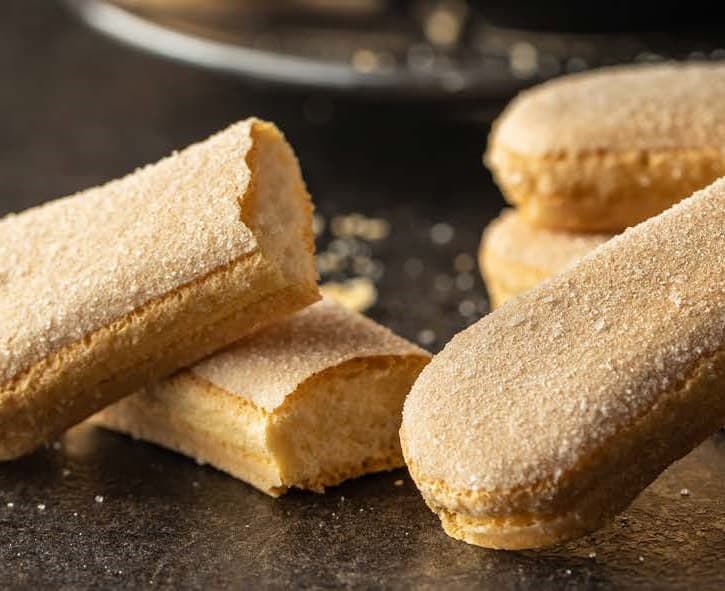 Long finger-shaped biscuits on a black countertop. One is snapped in half showing the airy centre and crumbs are on the surface of the bench it sits on.