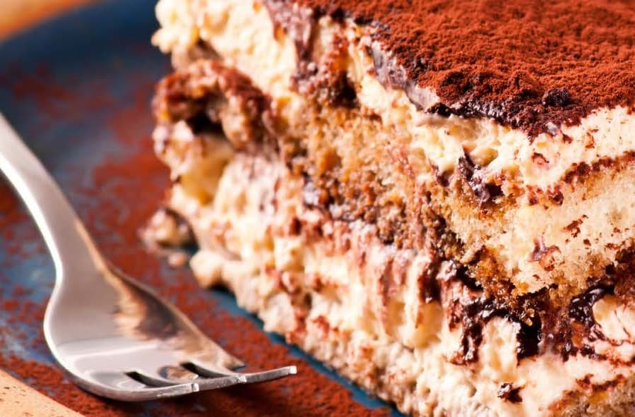 a fork lays alongside moist tiramisu, on a plate dusted with cocoa