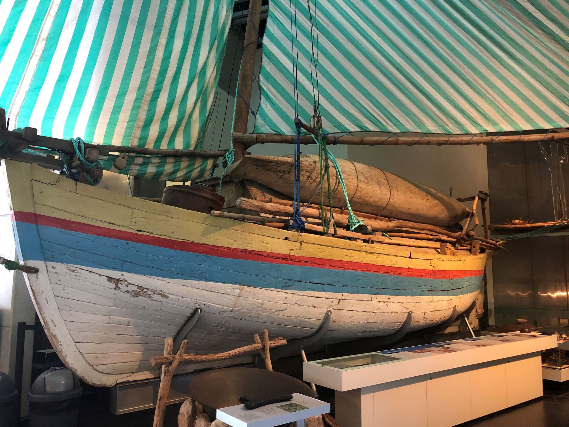 A small wooden sailing boat. The hull is painted white with yellow, red and blue stripes. The sails are striped blue and white.