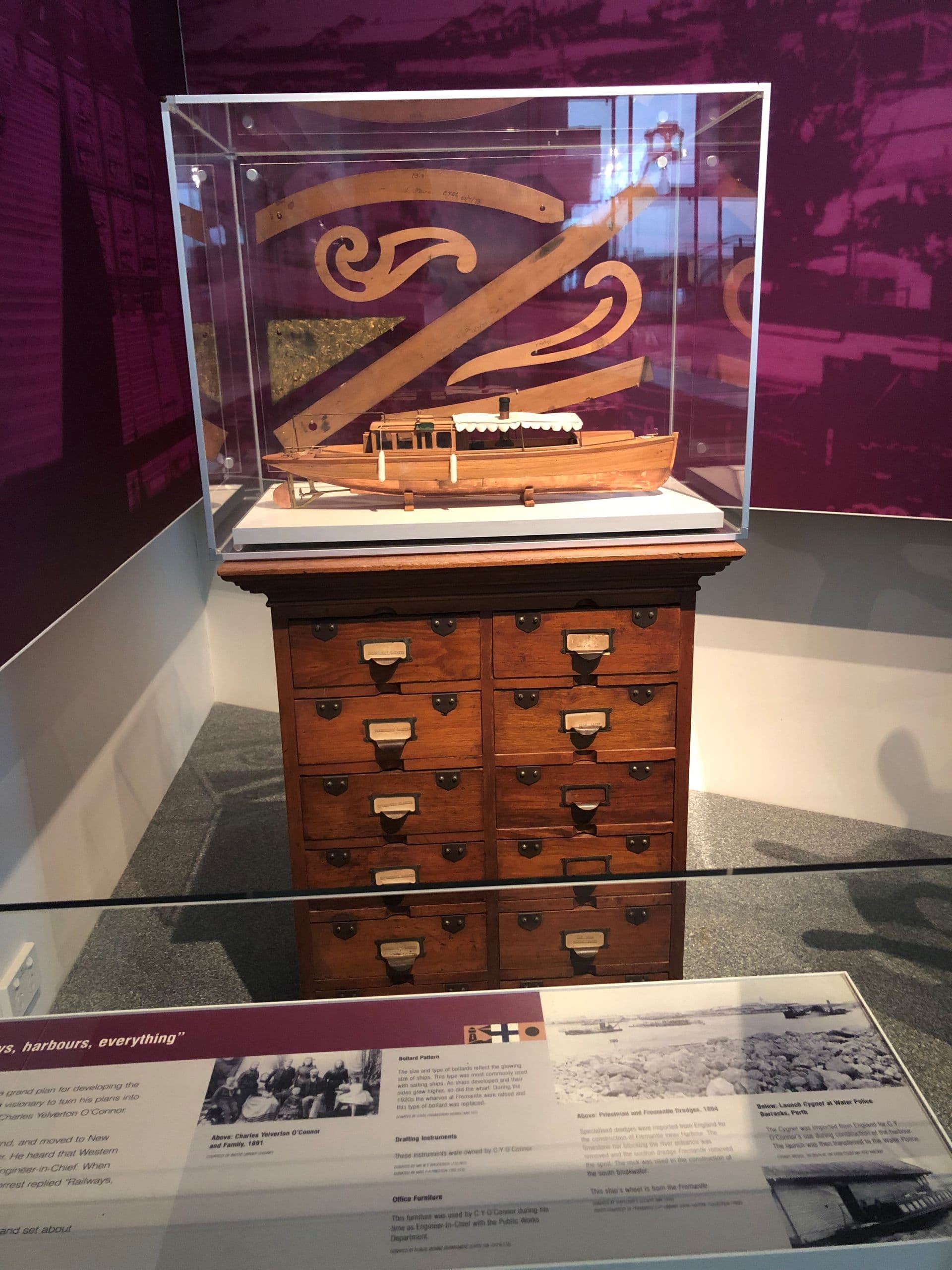 Behind a glass shield is a narrow dark wood cabinet with two columns of small drawers. On top is a glass display containing a wooden boat model.
