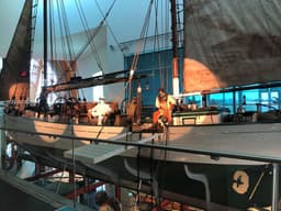 A large white and dark green sailing boat suspended next to the top floor. A mannequins in a diving suit sits  on the deck.