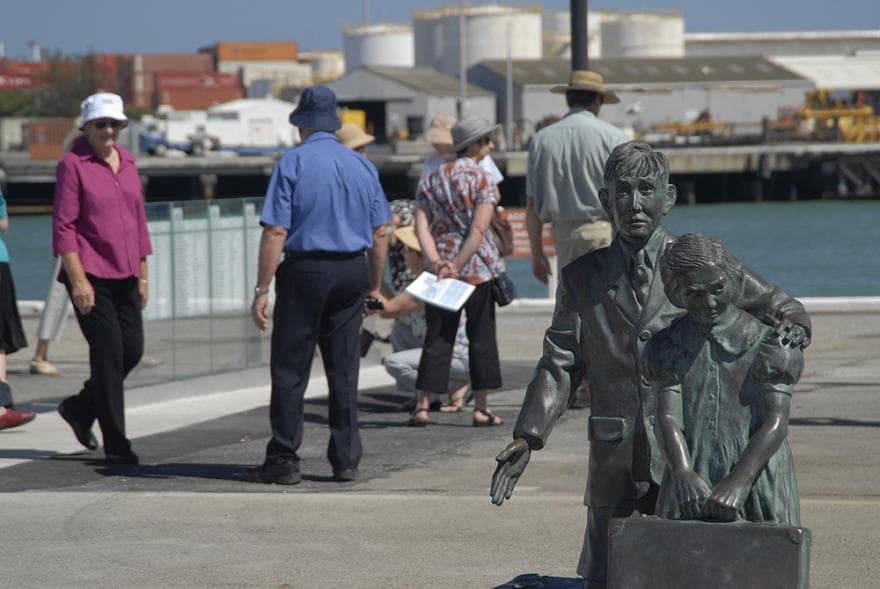 In the foreground is a statue of 2 small children with suitcases. In the background there are people wandering along the port with views of water and sheds behind them.