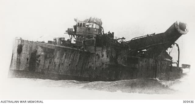 A grainy photo of a severely damaged metal ship. The ship's bridge is partly torn off and a tall funnel leans over the ship's side as if about to topple overboard.