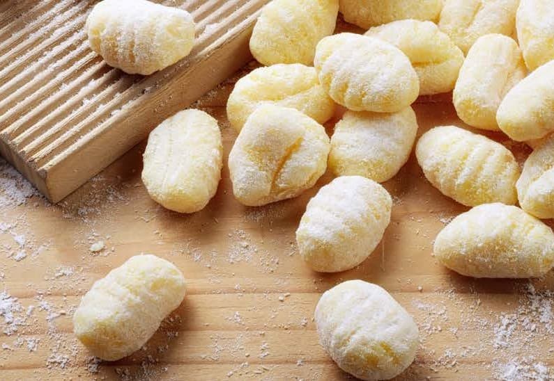 uncooked gnocchi sit on a floured wooden board. A wooden gnocchi roller is visible in the upper left corner if the image