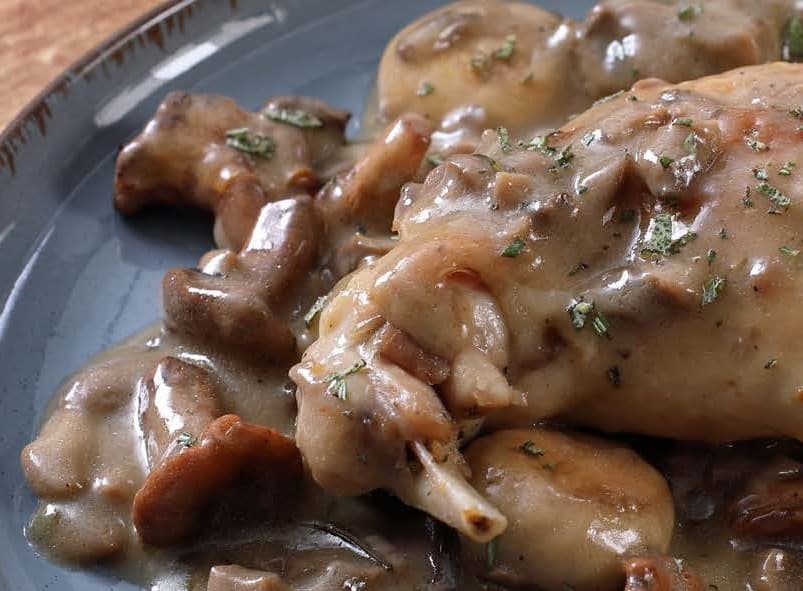 On a blue plate rests a joint of cooked rabbit, smothered in a glistening mushroom sauce, and garnished with finely chopped parsley.