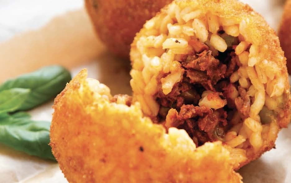 A ball of crumbed and fried rice, with a meat sauce, cheese and pea filling. It is cut open to show the centre and sits on some parchment baking paper on a wooden board with a basil leaf garnish