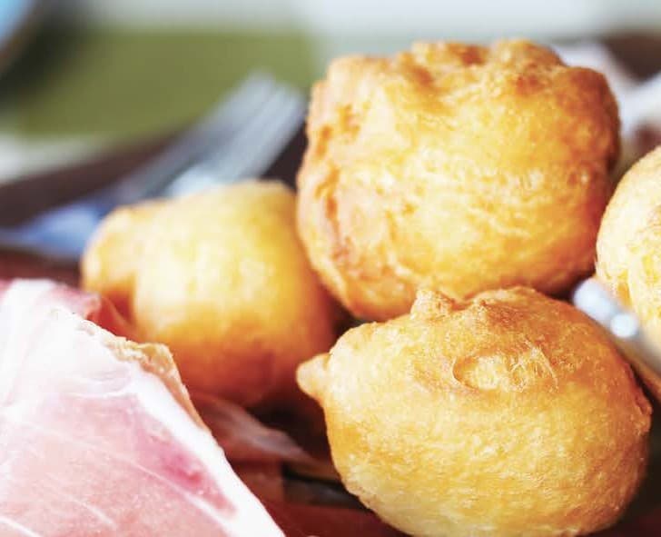 fried dough balls in a pile on a plate, next to some proscuitto