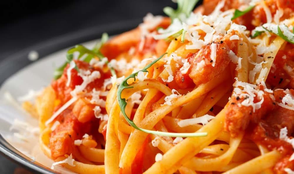 long strands of spaghetti pasta tossed through a red sauce and topped with grated cheese. It sits on a white plate with a black rim.