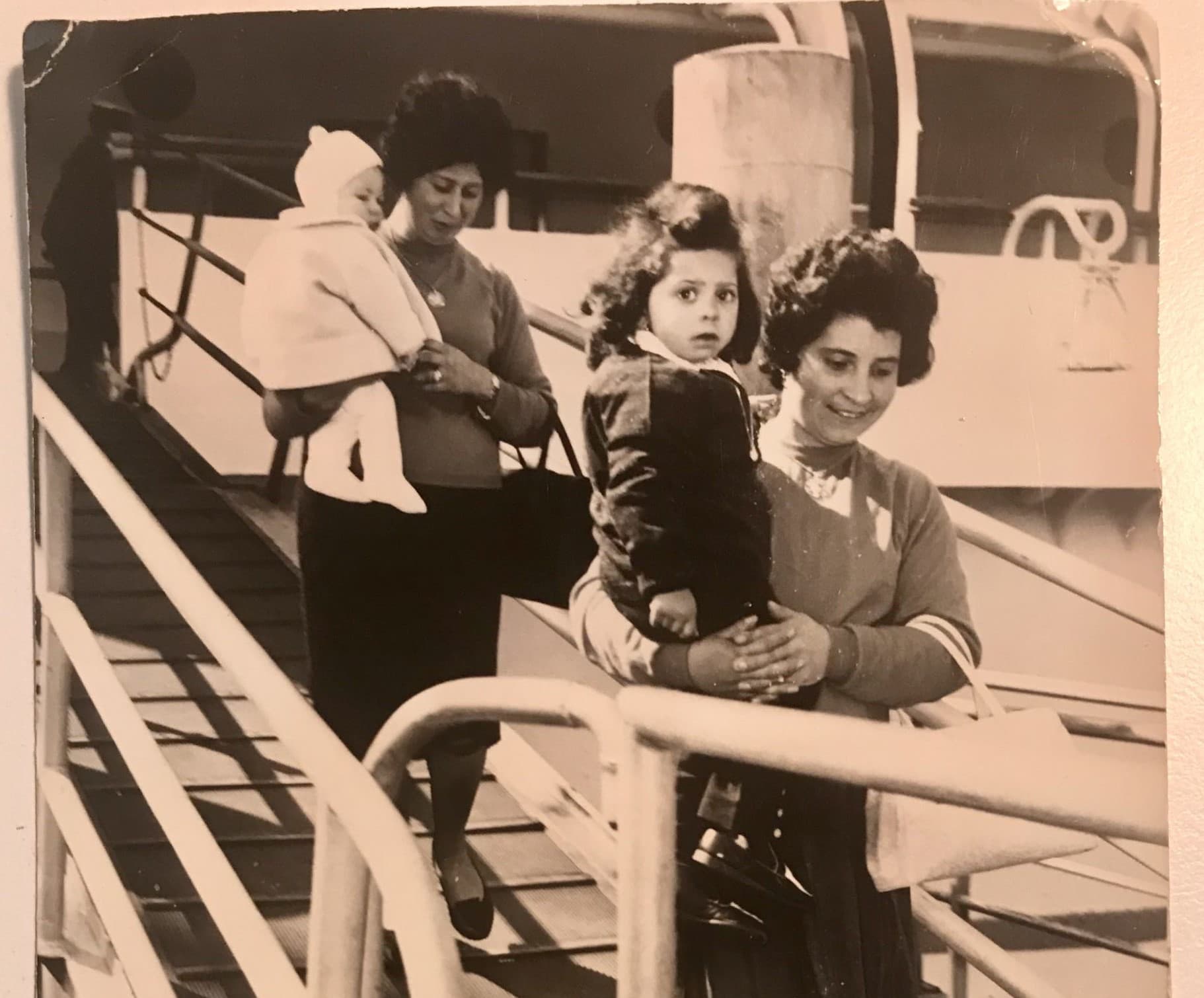 Two women holding small children walk down a gangway from a ship
